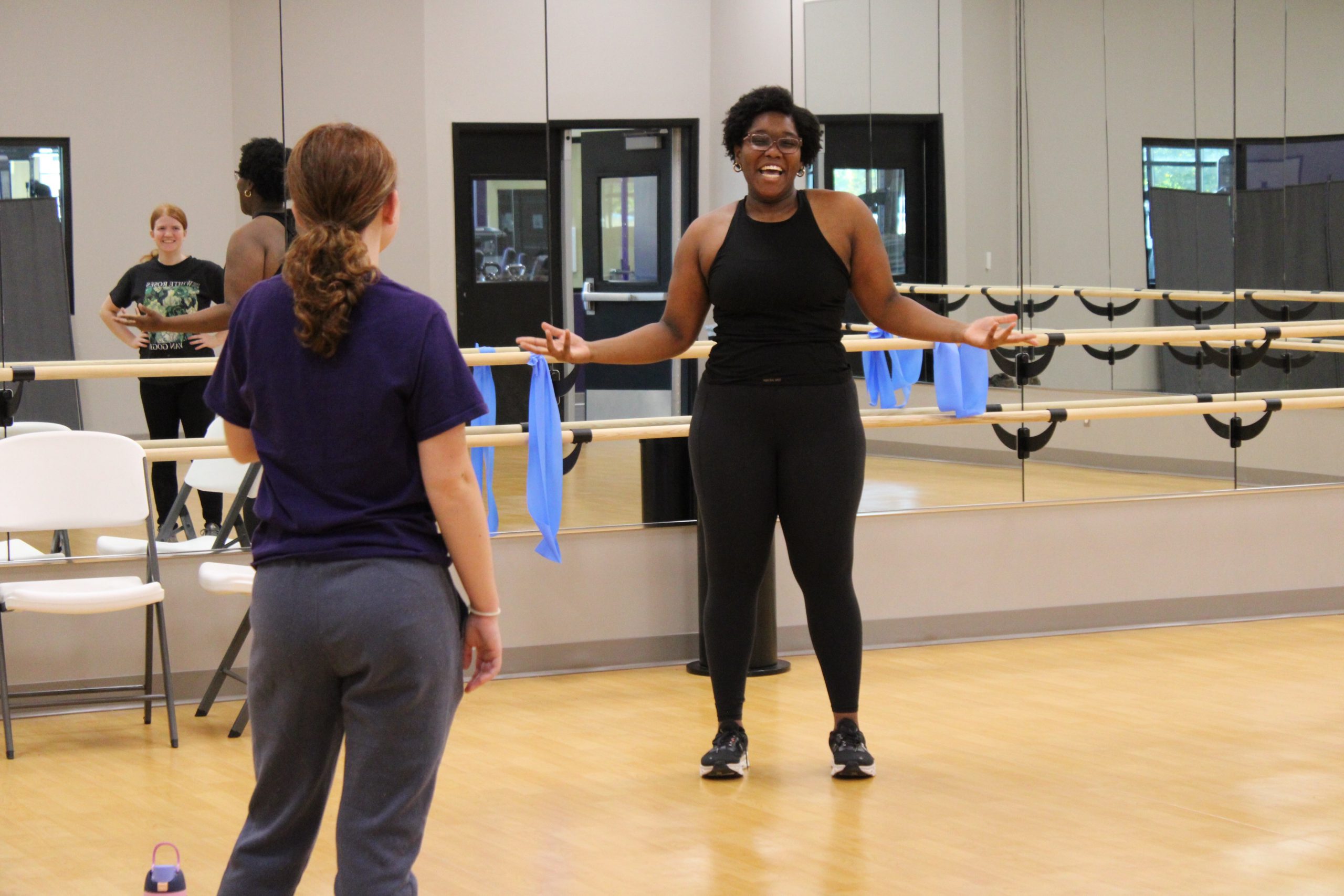 A young woman stands, arms outstretched and grinning, in front of a member of the class. The room is well-lit and surrounded by mirrors, reflecting another member of the group, smiling.