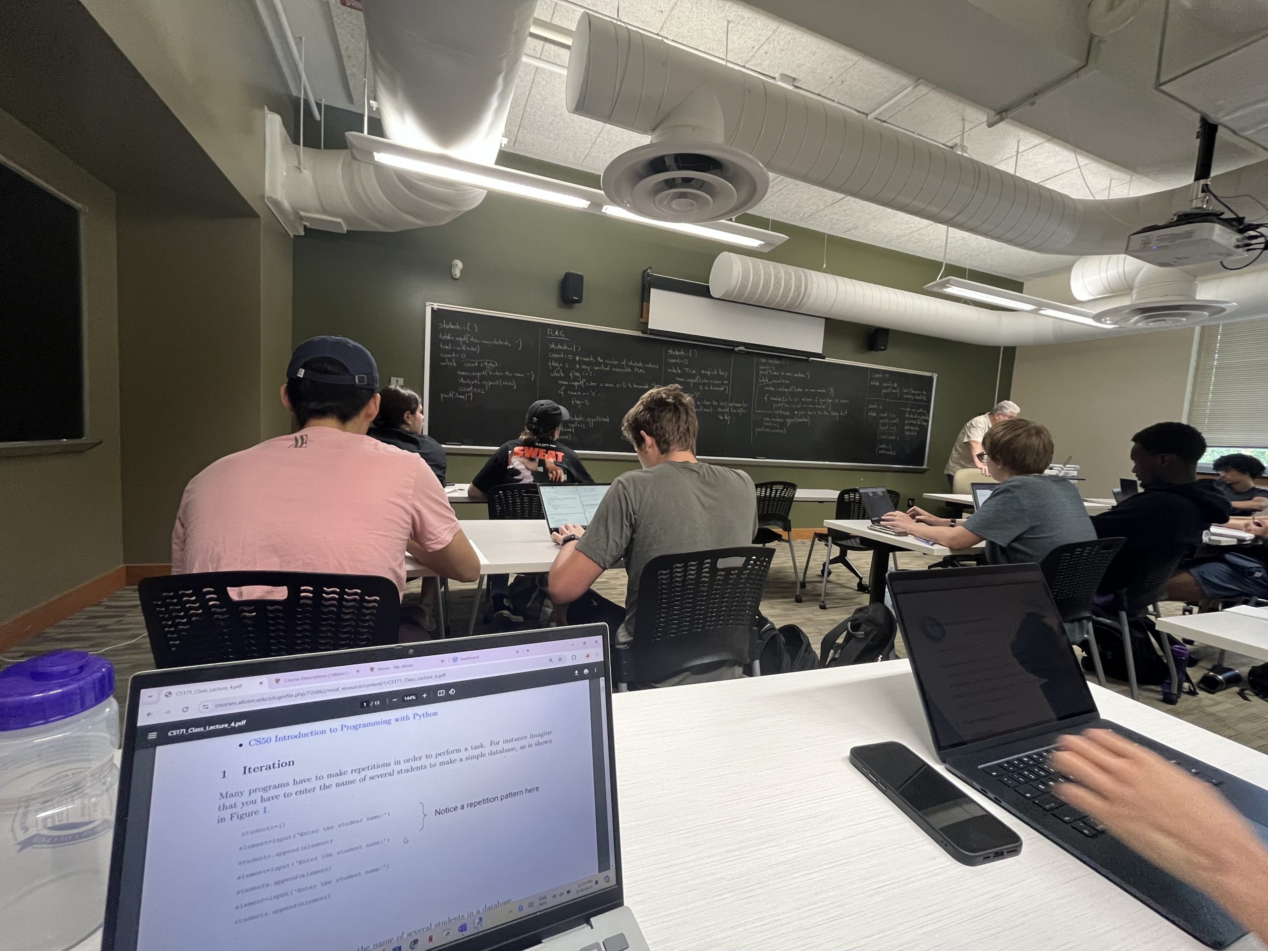 Students sit at desks, working on laptops, while the lecturer stands at the front of the classroom, explaining the concept of iteration on a blackboard. The classroom has rows of desks and a projector screen.