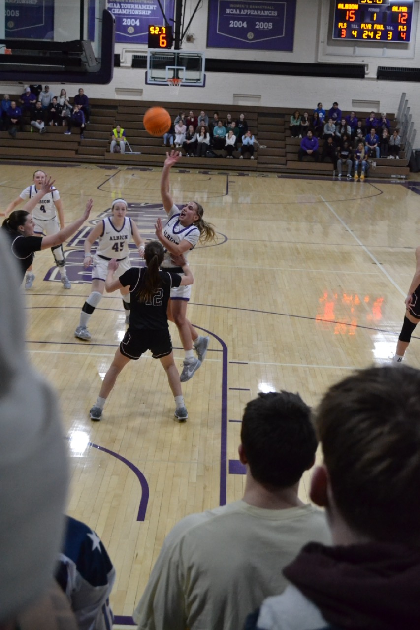 Basketball game action shot with a player in a white jersey jumping for a shot against a defender in a black jersey. A crowd watches from the bleachers on the other end of the court.